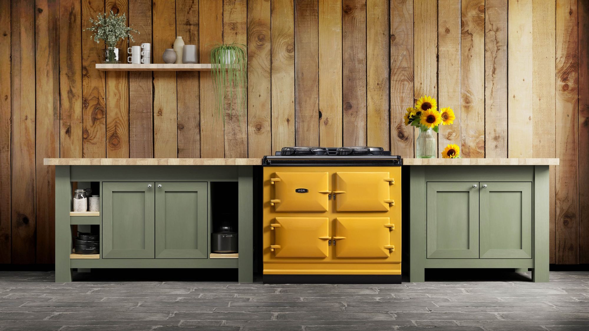 Cuisinière AGA de 100 cm coloris moutarde entre deux meubles vert sauge, mur en bois brut
