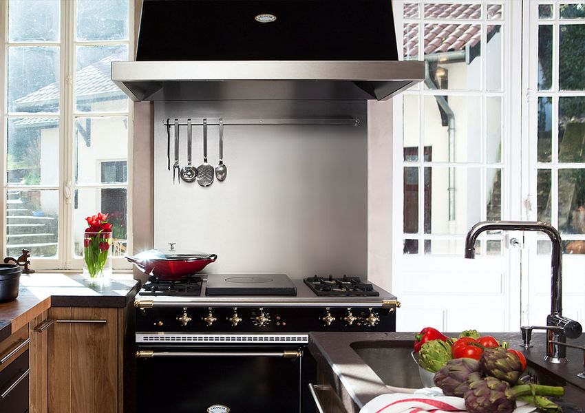 Piano de cuisson Lacanche Classic noir à finitions laiton, sous une hotte inox, dans une cuisine lumineuse