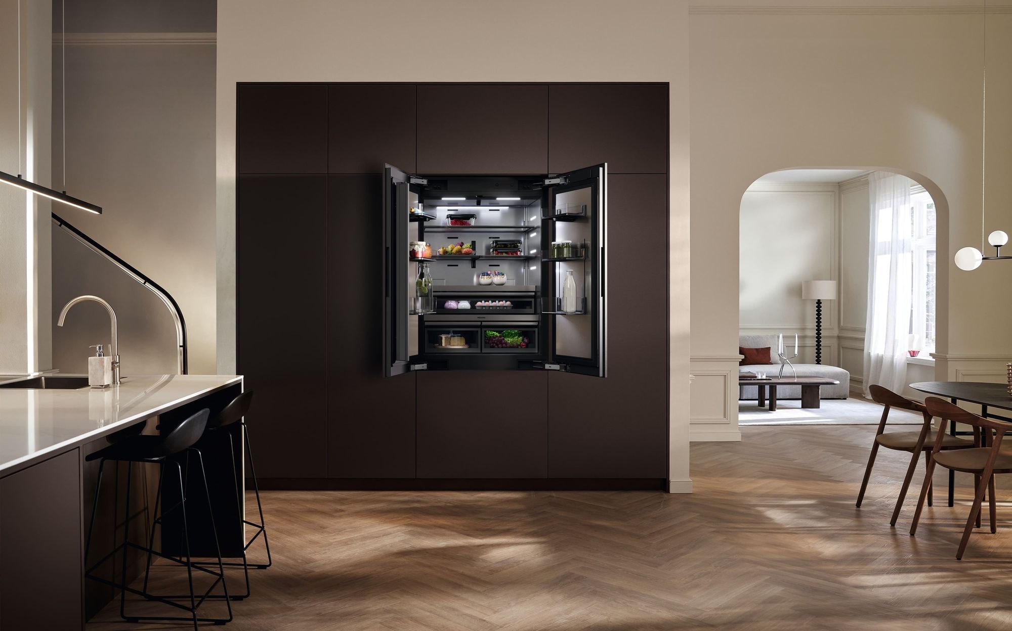 Réfrigérateur Miele MasterCool FrenchDoor ouvert dans une cuisine sombre au parquet à chevrons, ouverte sur la salle à manger
