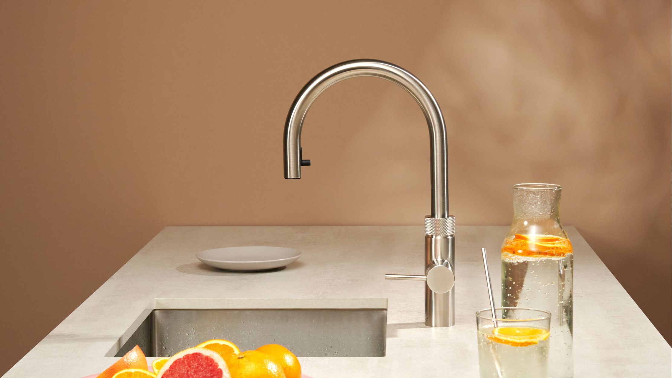 Robinet Quooker Flex Round inox au-dessus d’un évier sous plan, mur rose poudré et carafe d’eau aux agrumes